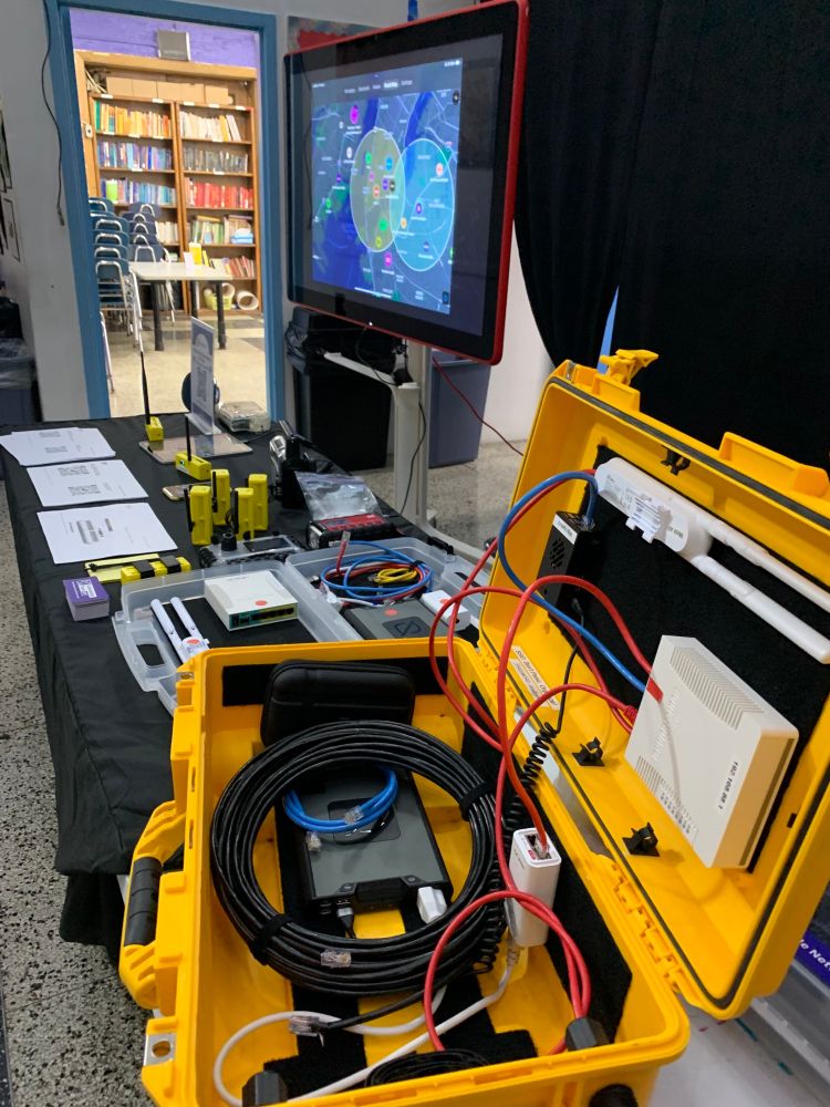 Table with black table cloth displaying informational sheets, Meshtastic/Lilygo devices in yellow cases and two PNKs, one in the yellow case open to display hardware. Behind is a screen showing other live meshtastic devices in the area