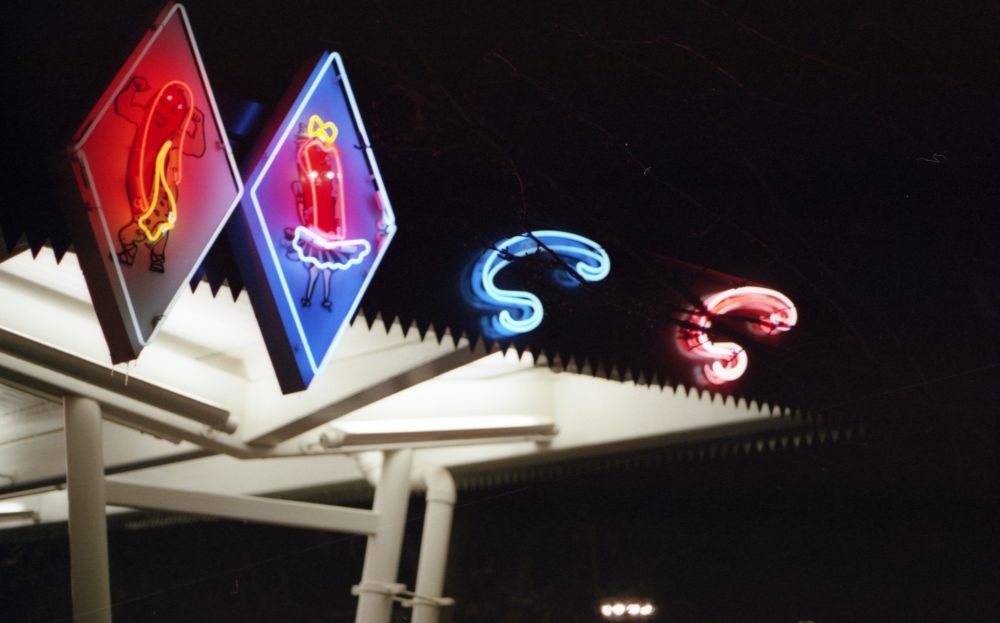 Two neon diamonds, one reddish pink one blue featuring the Superdawg mascots. The red diamond features the boy hot dog mascot flexing his muscles wearing an animal hide like an old timey circus bodybuilder. The blue diamond features the girl hot dog mascot who is wearing a short blue frilly skirt with a yellow bow on top. Next to it on the right are two neon Ss, one blue and one red. Underneath you can see the lights and fixtures for the drive in cover
