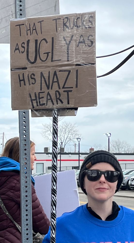 Person in sunglasses holding a hand lettered cardboard sign on a stick that reads “That truck’s as Ugly as his Nazi heart” 