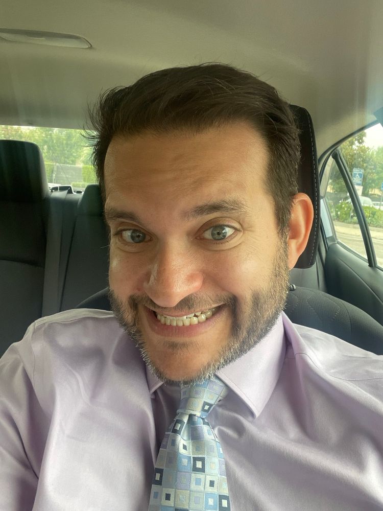 A guy with a beard smiling wearing a lavender shirt and a tie. 