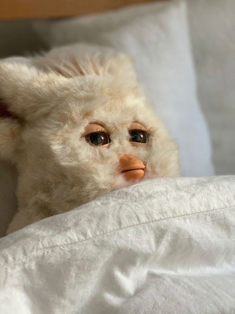 a white Furby lying in bed under a soft sheet, eyes half-open in a way that perfectly captures the look of someone who’s both tired and mildly regretting all of their life choices