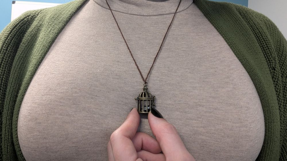 a woman in a tan turtleneck and green cardigan is holding between her thumb and forefinger against her chest a very tiny birdcage pendant with a very tiny person inside who's reading a newspaper and is probably totally comfortable in there, trust me.
