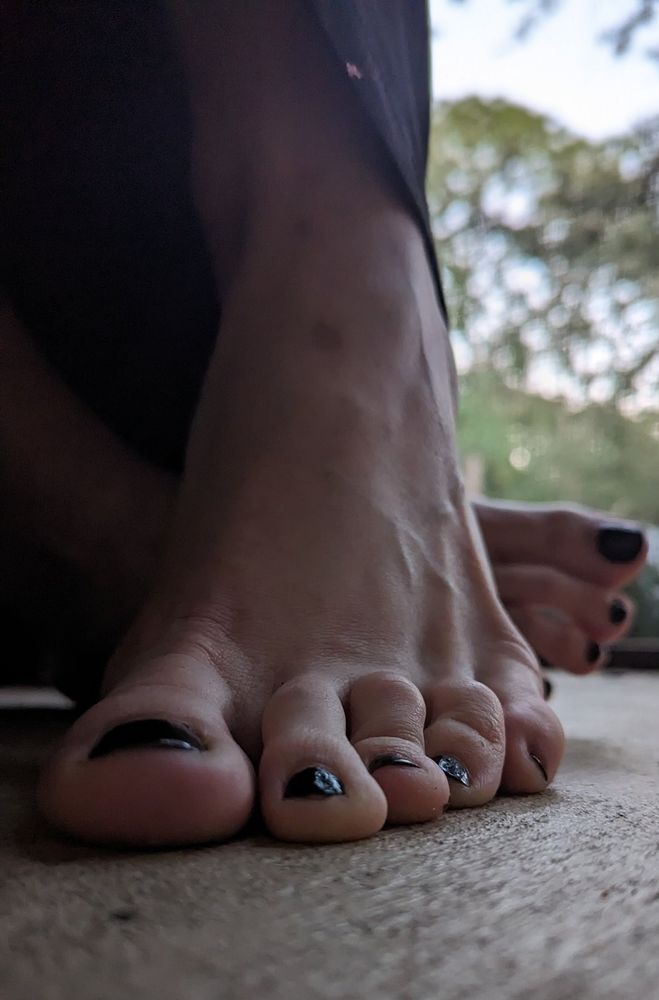the top of a woman's left foot faces the ground-level pov with the heel raised and the other foot resting behind it on its side as if the owner is sitting down and leaning over, casting shadows overhead. the nails are black.