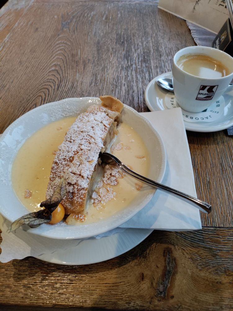 Ein Teller Apfelstrudel mit Vanillesauce und daneben eine Tasse Kaffee.