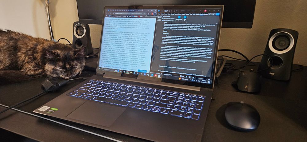 A medium haired tortoiseshell cat peacefully naps on a desk beside a laptop open to a manuscript draft and scientific paper being referenced.