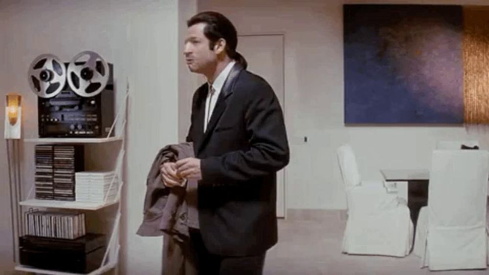 a man in a suit and tie is standing in a living room next to a shelf .