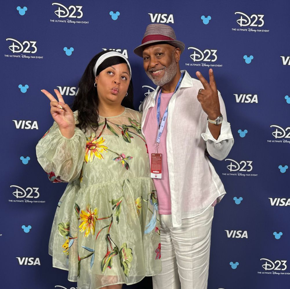 Gavyn wearing a green tulle dress with yellow, blue and green flowers embroidered all over it. She is next to her dad who’s wearing white linen suit pink shirt and straw fedora. They’re standing in front of. Disney D23 step and repeat holding up peace signs. 