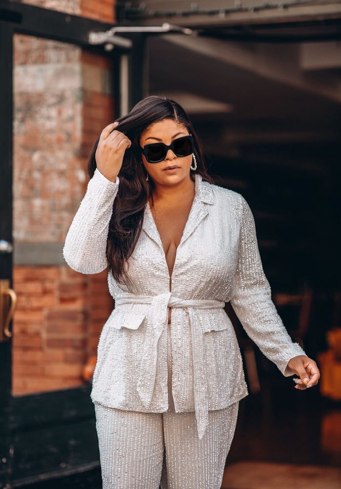 Gavyn from the knees up standing just outside of a restaurant door way wearing square dark sunglasses, an all white soft blazer covered in beads and sequins tied at the waist, showing a little cleavage, and matching pants 