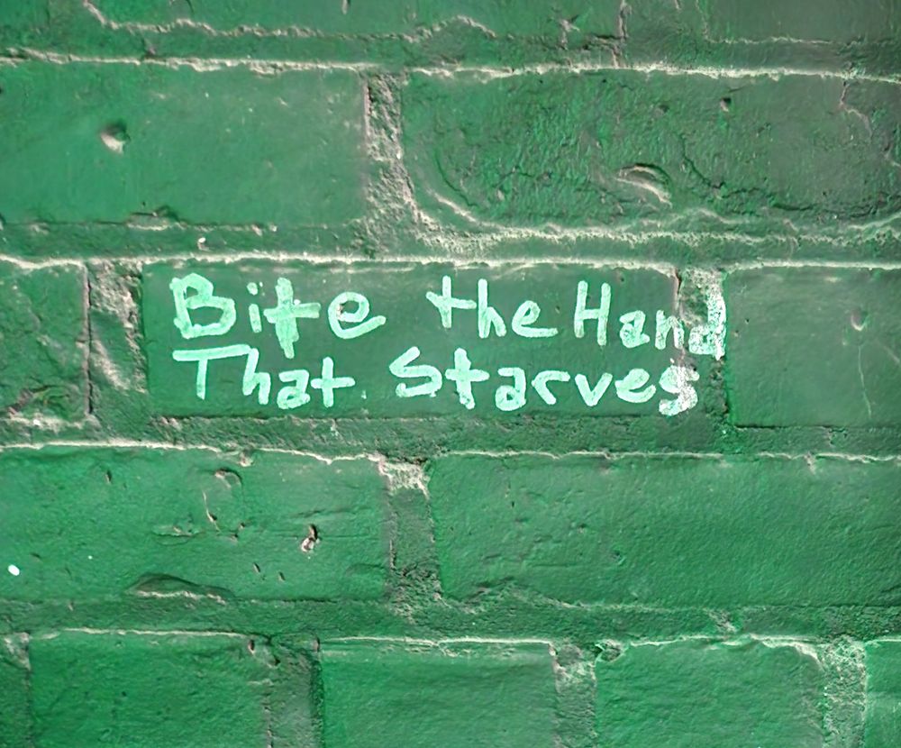 Graffiti on a brick wall in a nightclub that reads "Bite the Hand That Starves". A green glow illuminates the wall and text from the club lighting.