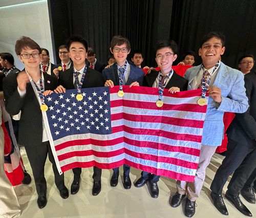 Five students wearing medals stand holding the American flag, smiling after a competition. They are dressed in formal attire, surrounded by other participants in the background.