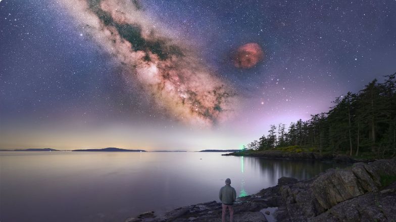 Person standing on rocky shore under a vivid Milky Way stretching across the night sky.