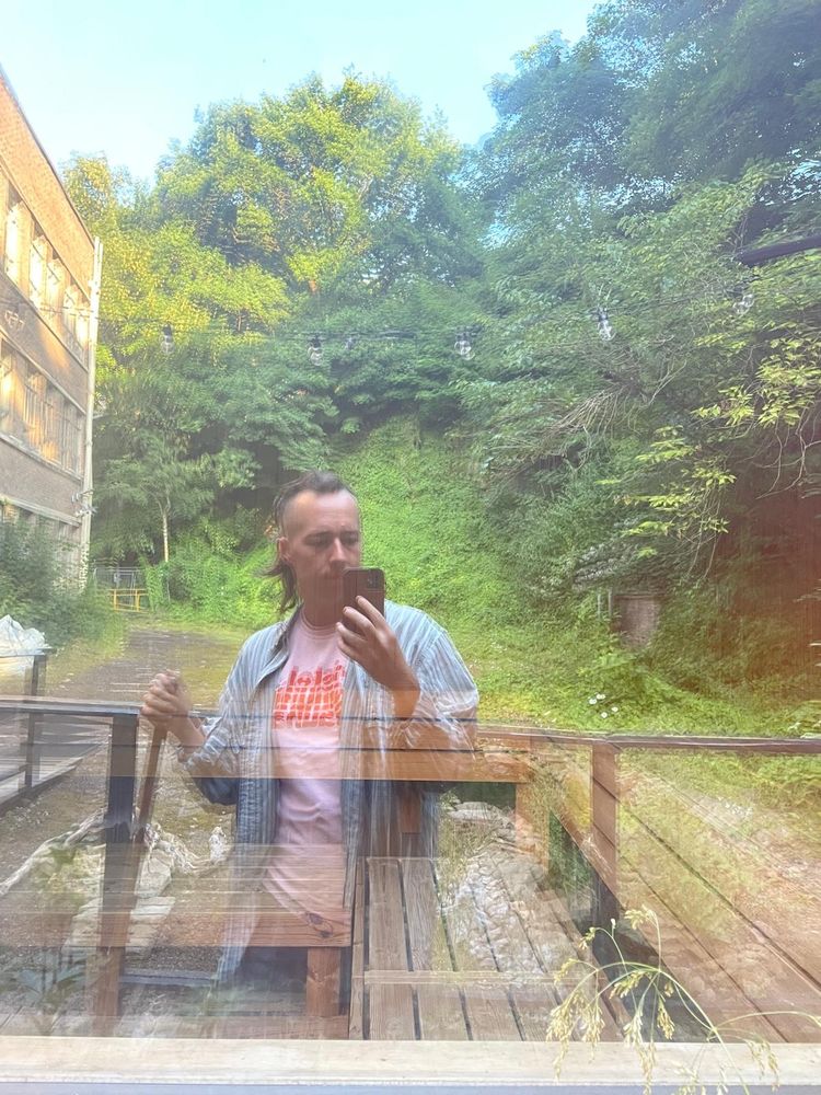 Joe is wearing a pink t-shirt and a blue overshirt. They appear as a reflection in a window. Through the window, wooden benching can be seen. Elsewhere in the reflection there are lots of verdant trees, bushes, invasive species and a car park.
