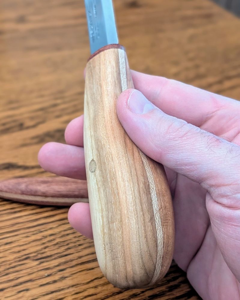 A large sloyd knife with a cherry handle held lightly in a man's hand. The tip of the blade is out of frame.