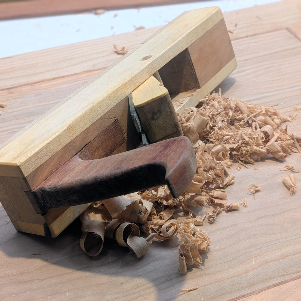 A wooden plane sitting on its side, on top of a cherry wood panel, with plane shavings spilling out.