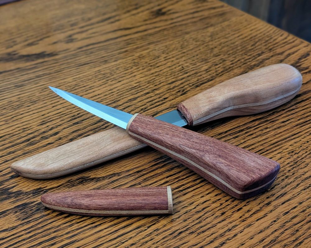 2 knives with wooden blades, crossed. On top is a smaller knife with a red African rosewood handle. On the bottom is a larger knife with a cherry handle and matching sheath