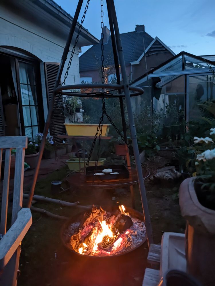 An outdoor scene at dusk featuring a large, dark metal tripod structure with a fire pit at its base. Above the fire, a grill and pots are suspended. 

A house with shuttered windows and a greenhouse are visible in the background.