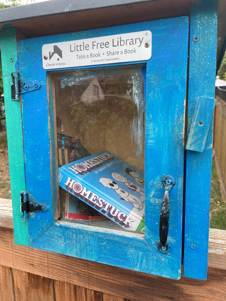 The Homestuck graphic novel inside a free library. A plaque at the top of the library reads “take a book leave a book”