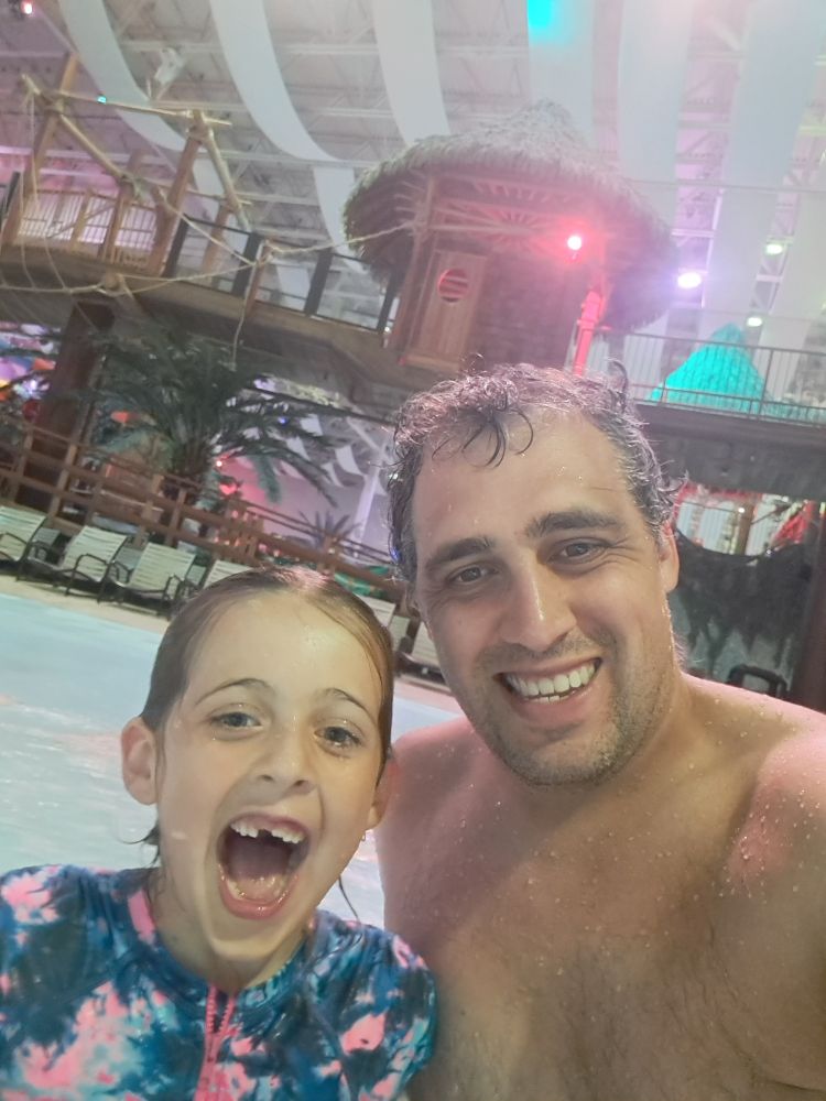 Handsome young man who looks physically too young to be a father but you can tell he has deep emotional maturity poses with his daughter who for some reason took all photos with her mouth fully ajar at the wave pool moments before the park closed and the water works moved from the bora bora slides to her eyes suck it john robb