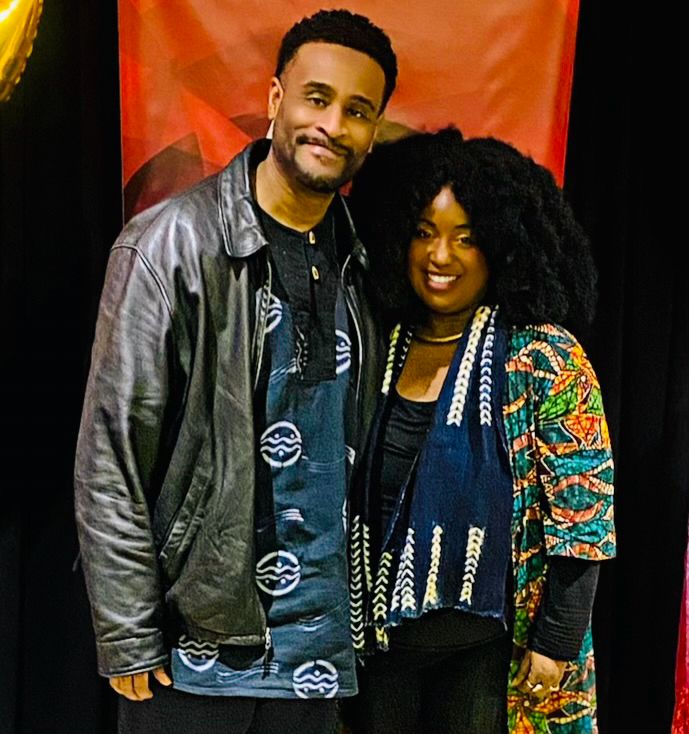 Sheree and her husband, smiling before the red Keeper of the Cultural Flame Award backdrop / step-and-repeat vertical banner