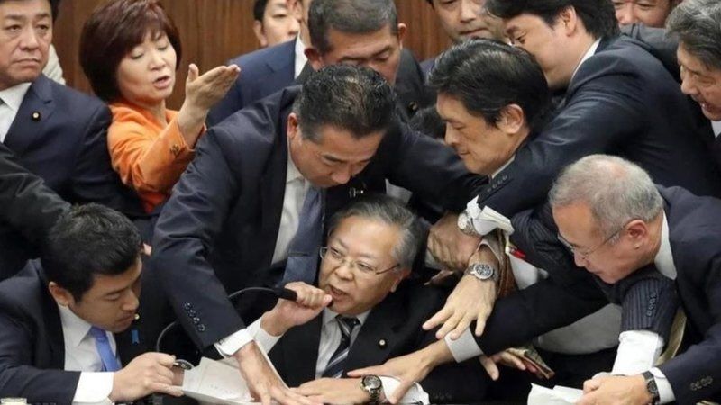 Several people in suits reach towards a man with glasses sitting at a table, holding a microphone. The latter speaks into the microphone, while the former attempt to stop him.