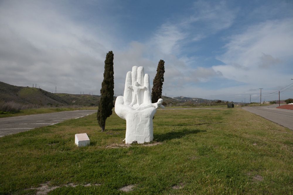 Sculpture in white of a 12 foot hand coming vertically out of the ground with palm horizontal and fingers vertical. On the palm are two children
