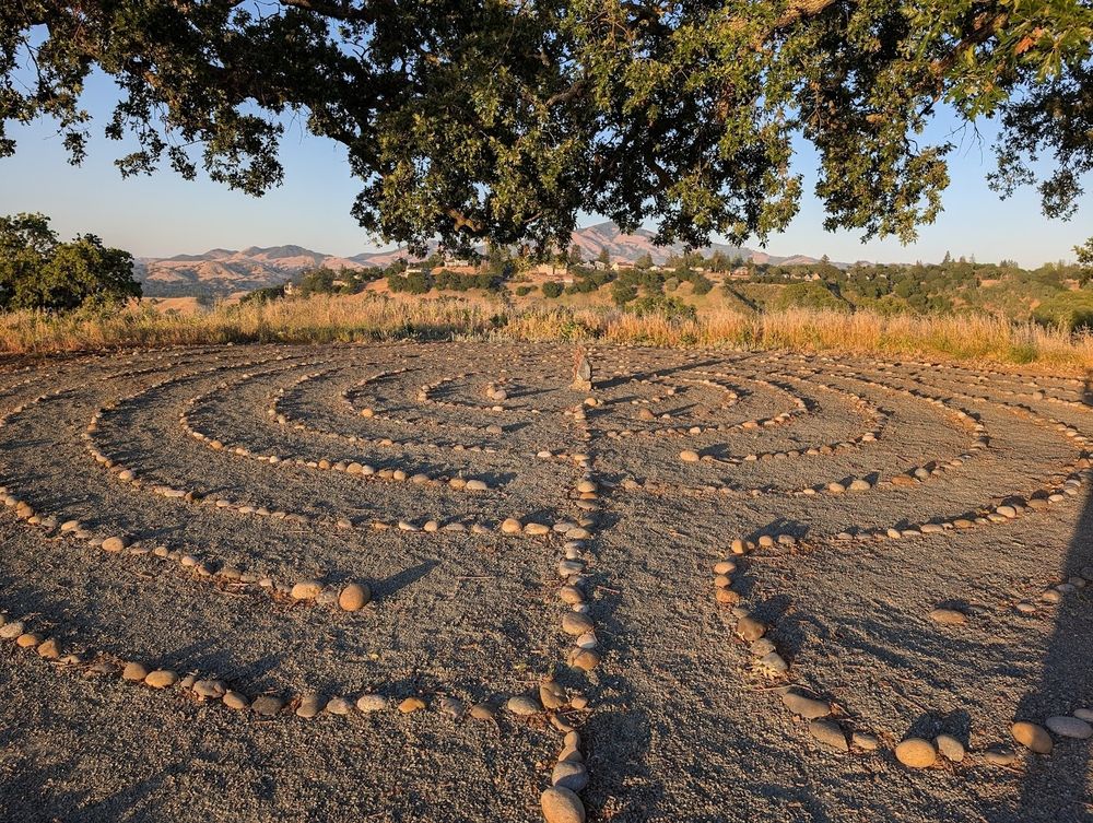 Photo of a rock labyrinth at sunset at the top of a hill that a local rock club created about 10 years ago.  A large white oak tree overlooks it. Mount Diablo is barely visible in the background. 