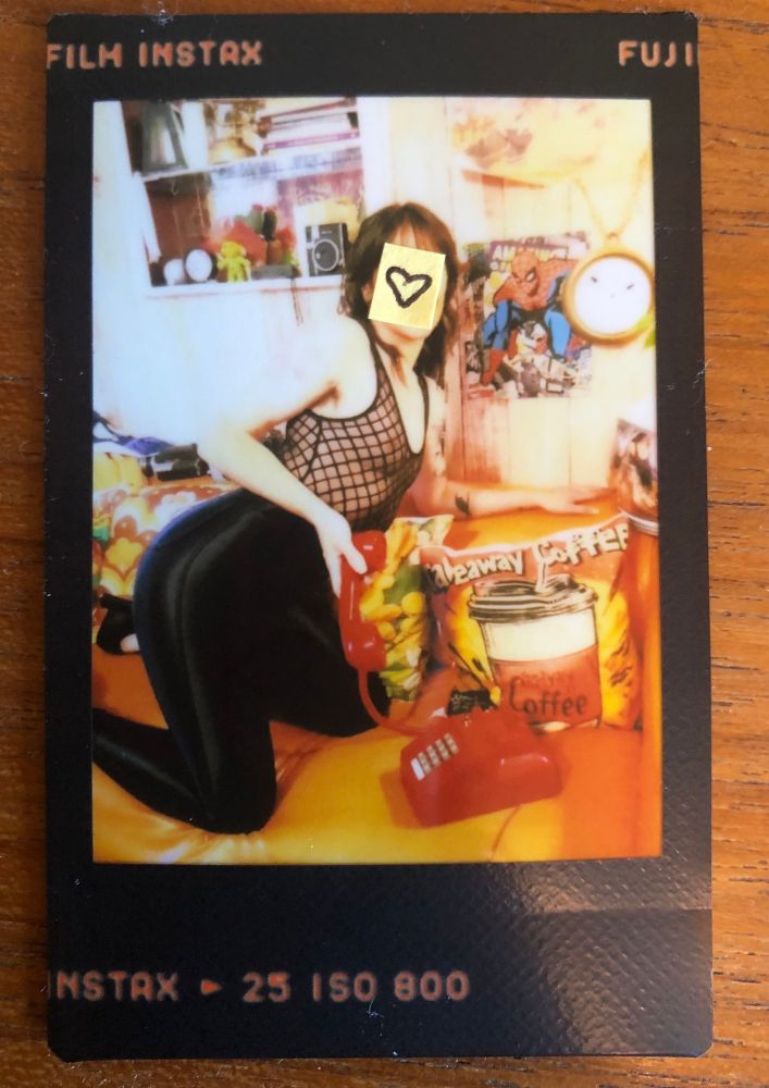 a polaroid picture of sadie in tight black hot pants, mesh black bodysuit, and heels. she bends over an orange couch in a 70s living room while talking on a retro red landline phone. 