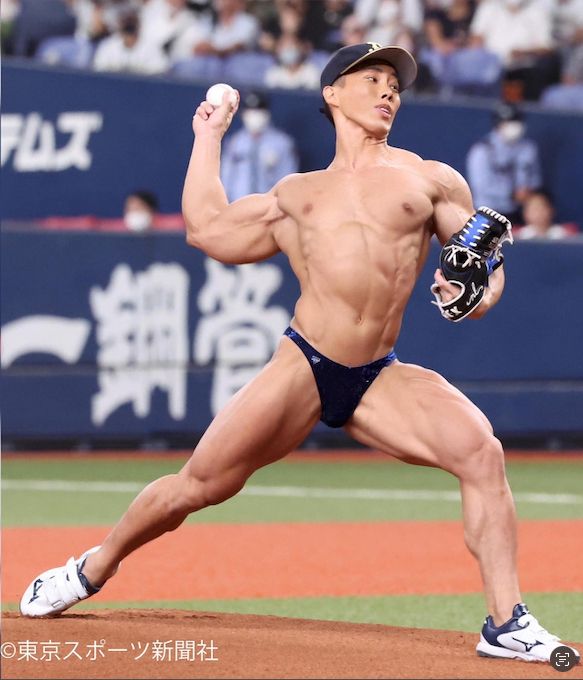 A photo of japanese bodybuilder Naotaka Yokokawa throwing an opening pitch during a baseball game. He's wearing a blue, velvet speed, sneakers, and a baseball cap. He has a baseball catcher's mitt loosely held on one hand. 