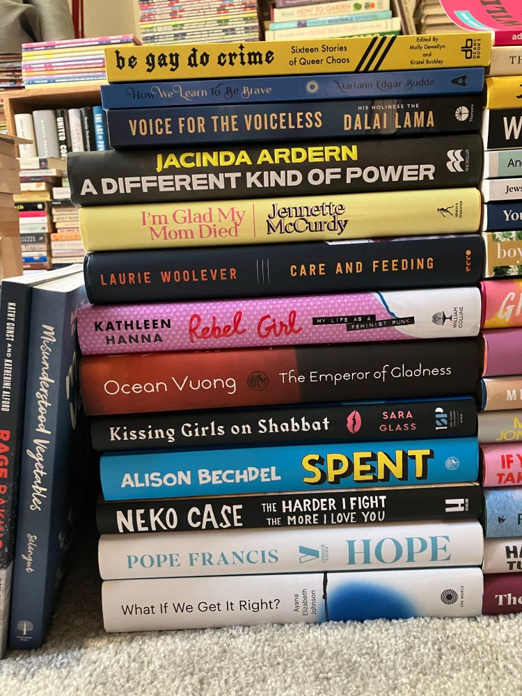 A photo of a stack of 13 books, a mix of fiction and non-fiction, mostly new hardbacks, including (from bottom of stack to top) ‘What If We Get It Right?’ by Ayana Elizabeth Johnson, ‘Hope’ by (the late) Pope Francis, ‘The Harder I Fight The More I Love You’ by Neko Case, ‘Spent’ by Alison Bechdel, ‘Kissing Girls On Shabbat’ by Sara Glass, ‘The Emperor of Gladness’ by Ocean Vuong, ‘Rebel Girl’ by Kathleen Hanna, ‘Care and Feeding’ by Laurie Woolever, ‘I’m Glad My Mom Died’ by Jennette McCurdy, ‘A Different Kind of Power’ by Jacinda Ardern, ‘Voice for the Voiceless’ (paperback) by his holiness the Dalai Lama, ‘How We Learn to Be Brave’ (paperback) no by Mariann Edgar Budde and ‘Be Gay Do Crime: Sixteen Stories of Queer Chaos’ (paperback) edited by Molly Llewellyn and Kristel Buckley. 
Also pictured (leaning against the book stack, almost out of shot) are the books ‘Misunderstood Vegetables’ by Becky Selengut and ‘Rage Baking’ by Katherine Alford and Kathy Gunst.
