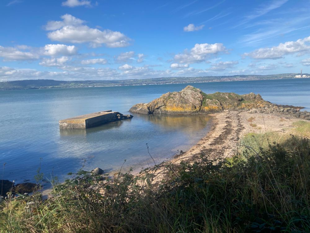 A natural curve of rocks above the waterline is enhanced by a man made concrete jetty, part of the defences left over from World War II when Belfast Lough was a potential Nazi target. The water is tranquil all the way to the farther shore, reflecting blue sky and white clouds in the upper third of the image. At the bottom, shoreline vegetation adds a touch of green, and thick shrubs out of shot cast a deep shadow, making the colours of rock, sand, water and sky even more intense. I'm pleased with this photo but it conveys only a fraction of the tranquillity of the moment. 