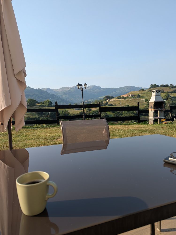 Foto desde una silla en la terraza del apartamento (a ras de suelo) de Cantabria, con mi café sobre la mesa y el prado de fondo.