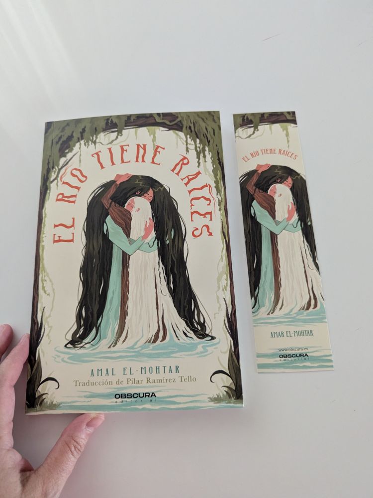 Foto del libro y el marcapáginas sobre una mesa blanca, con mi mano al lado. Se me da fatal describir ilustraciones, pero se ve a dos figuras abrazadas. La más baja tiene una larga melena blanca que se funde con el agua que tienen bajo ellas. La más alta se inclina sobre la primera y forma un arco con su pelo verde oscuro, casi negro, que la cubre por completo. Sobre ellas está el título en rojo, también formando un arco. Y, sobre el título, un arco de ramas y hojas que crea un pequeño marco que baja por los bordes de la cubierta. Debajo de la ilustración aparecen los nombres de la autora, la traductora y la editorial.