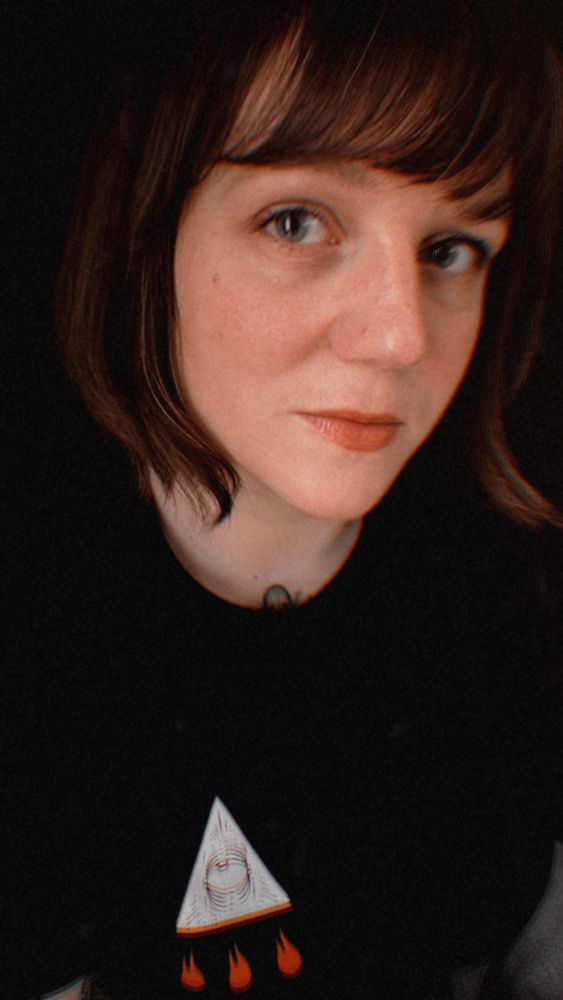 Photo de Agathe Boissonnot alias La Satanée portant un t-shirt noir avec un symbole représentant un œil dans une pyramide et 3 petites flammes en dessous sur la poitrine.