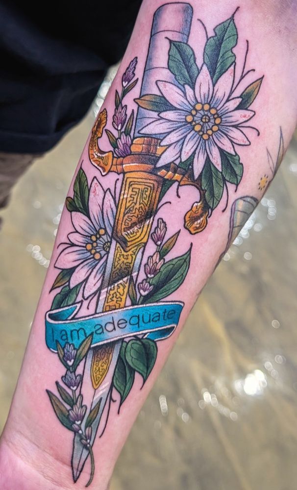 Dagger tattoo with edelweiss and lavender flowers and I am adequate quote written across the bottom on a real ribbon