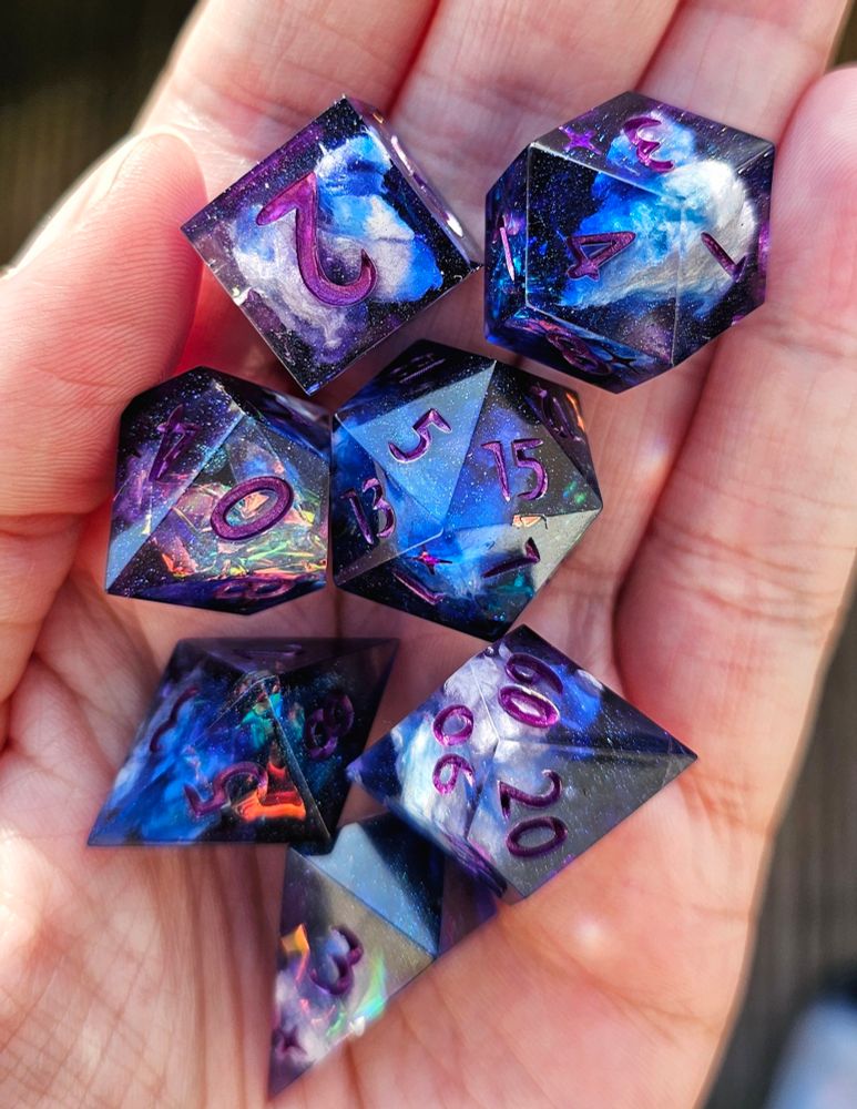 A 7 piece dice set on a hand. It's inked in purple. It's blue and purple with holo foil and clouds inside.