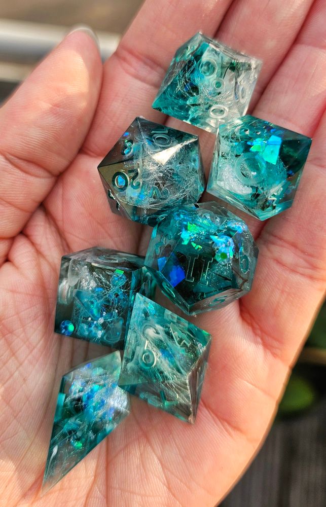 A set of raw dice (unsanded, unpolished, not inked) on a hand. They are turquoise/ greenish with green flowers, fabric, holo and white swirls inside.