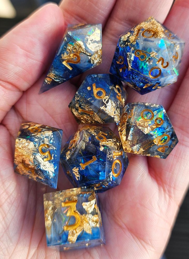 A 7 piece dice set on a hand. It's inked in gold. It's blue and clear with holo and gold foil and wire inside.