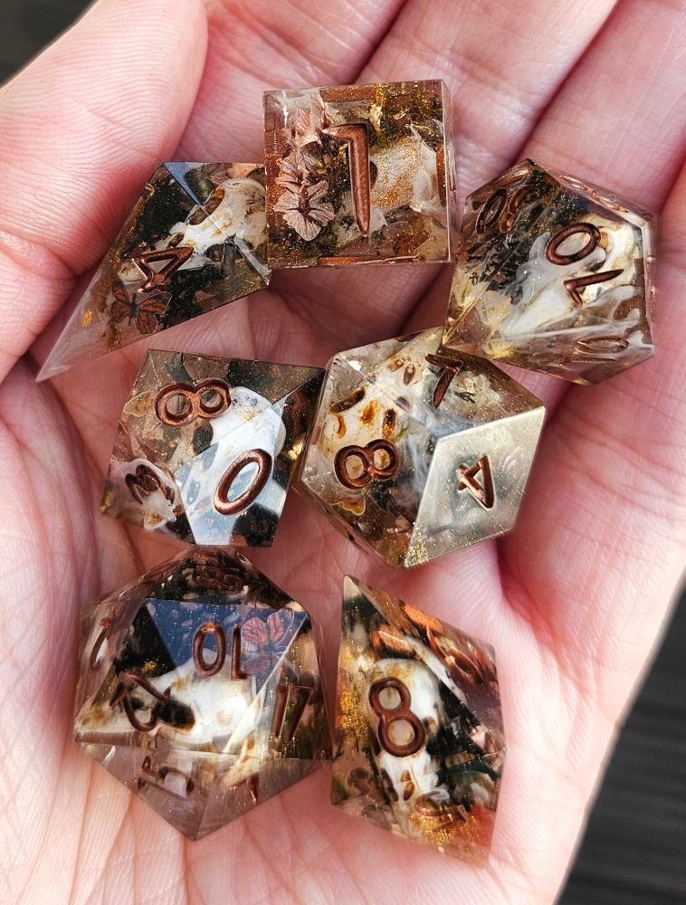A 7 piece dice set on a hand. It's inked in bronze. It's clear with hold glitter/ powder,  flowers and fake raven skulls inside. Also white swirls and moss.