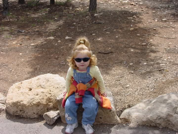 A toddler Raine sits on a rock with sunglasses on, red hair in a radish top ponytail. A green long sleeve under overalls, with butterflies on the ankles and a red, orange and green sweater tied around their waist. They’re rocking tiny white kicks and looking right at the camera 