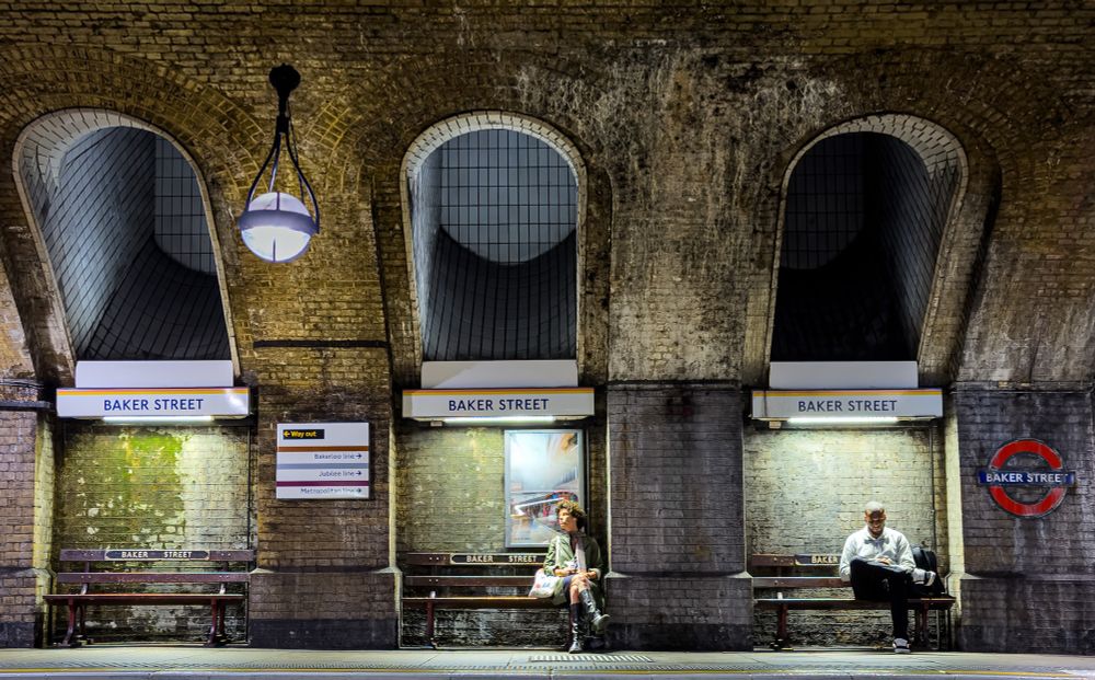 Two passengers sit on separate benches on one of the historic platforms at Baker Street Station for the London Underground. The Baker Street roundel is at the right, and the signage indicates the platform serves the Circle and Hammersmith and City Lines. 