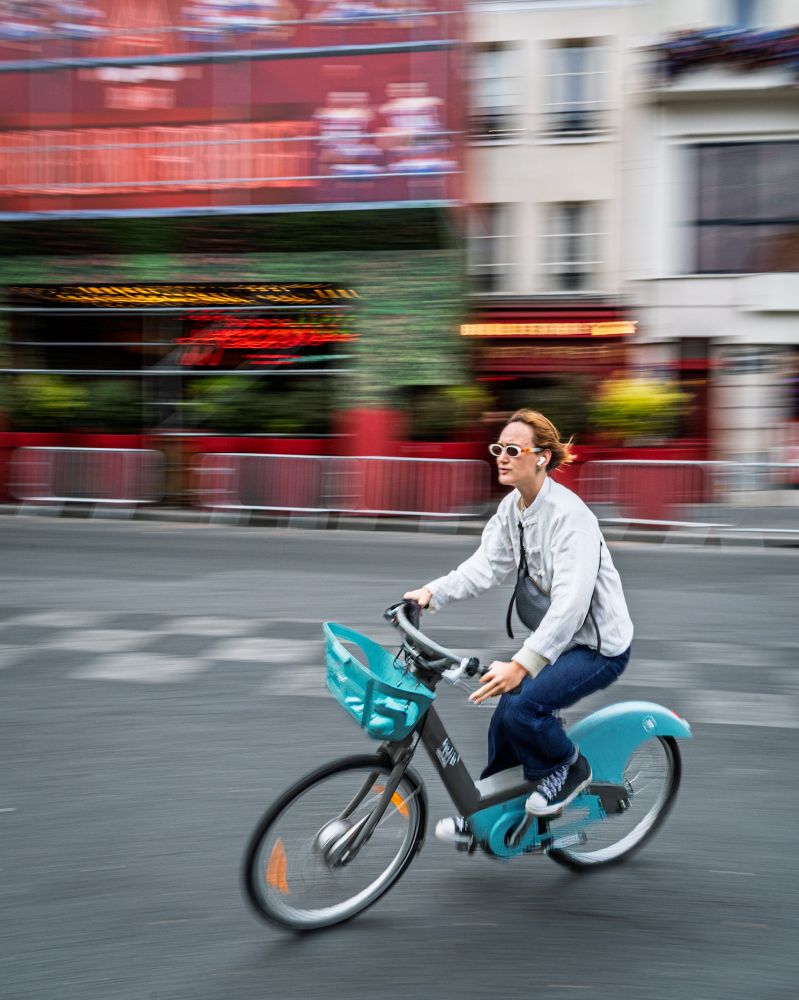 A woman on a teal bicycle rides in front of the Moulin Rouge in Paris. The woman is wearing distinctive sunglasses. The photographer has panned with the woman so she is mostly sharp while the background reflects motion blur. 