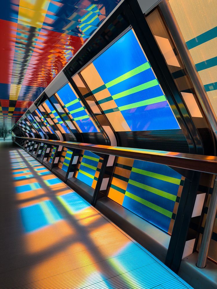 The interior of a modern, covered bridge. The walls are transparent and casting the colors of the walls on the floor as the light streams through. 