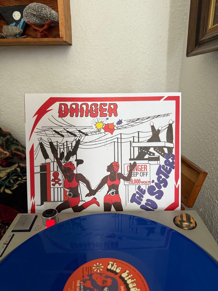 Blue record on a turntable with an album cover behind. There are red lightning bolts as a border and then a cartoon of the Lijadu Sistwrs in red outfits dancing around a power station. At the top in electrical red lettering it says DANGER