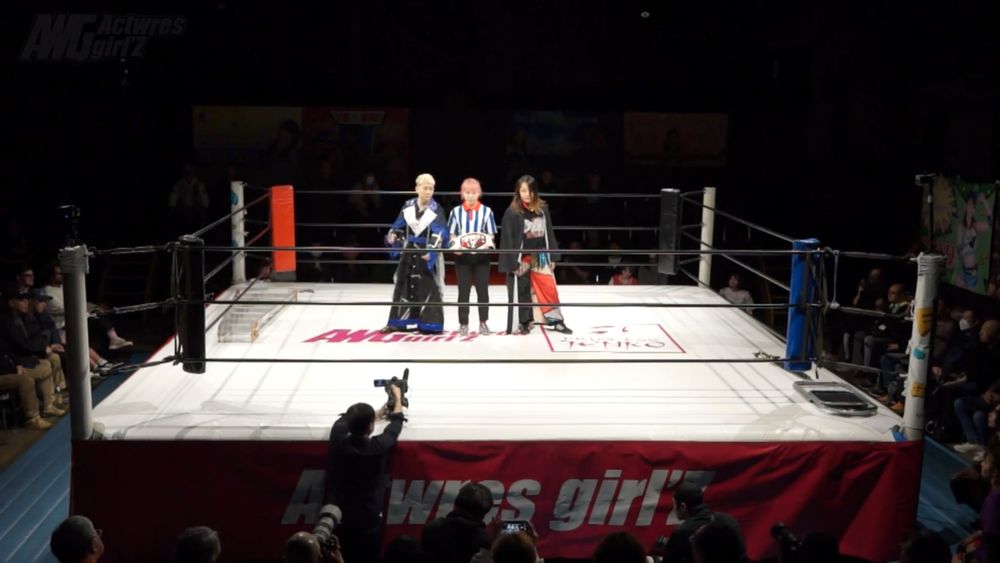 A distance pic of the ring. From L to R: MARU, the ref holding the KORE title belt, and Kyoka Iwai. There is a ladder in one corner and a chair in the other.