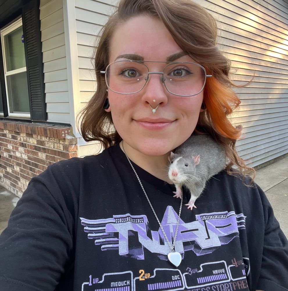 a femme presenting person with glasses, a septum piercing, and a slight smile, standing outside with a rat on their shoulder. The rat has blue-gray fur, a pink nose, and a distinctive white lightning bolt pattern on its face. The person is wearing the same black TWRP band shirt with a purple graphic. There is a natural glow from behind.