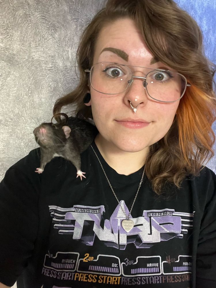 A femme presenting person with glasses and a septum piercing, smiling without teeth while a rat sits their shoulder. The rat has black fur, dumbo ears, and a white belly. The person is wearing a black TWRP band shirt with purple graphics.