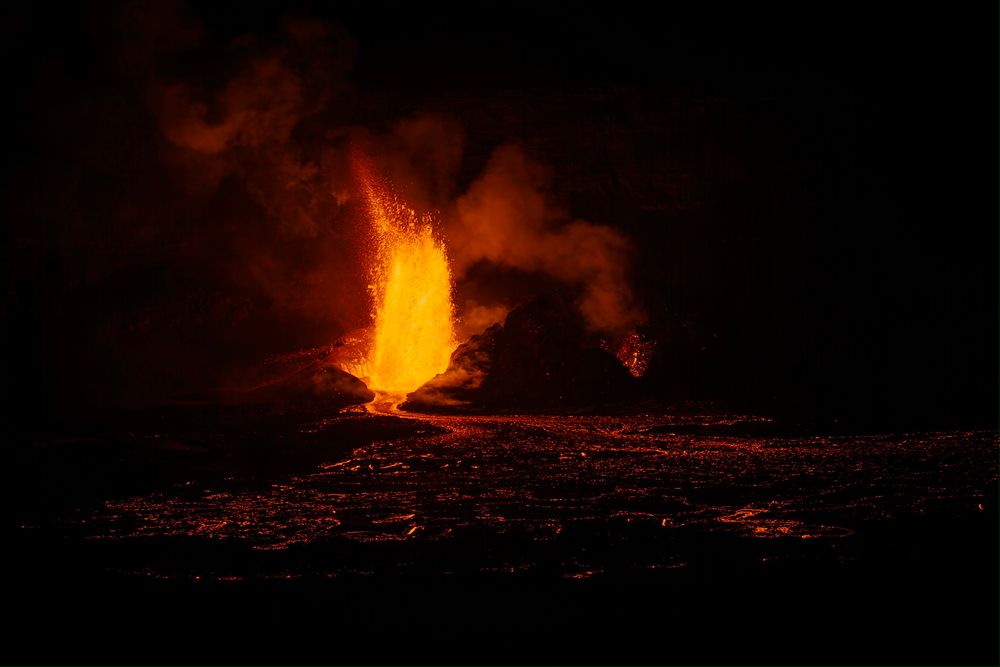 Wider shot of fountain of lava shooting upwards at night about 400-600 feet. There’s a small river of lava flowing in front of it. It flows into a sea of glowing magma. 
