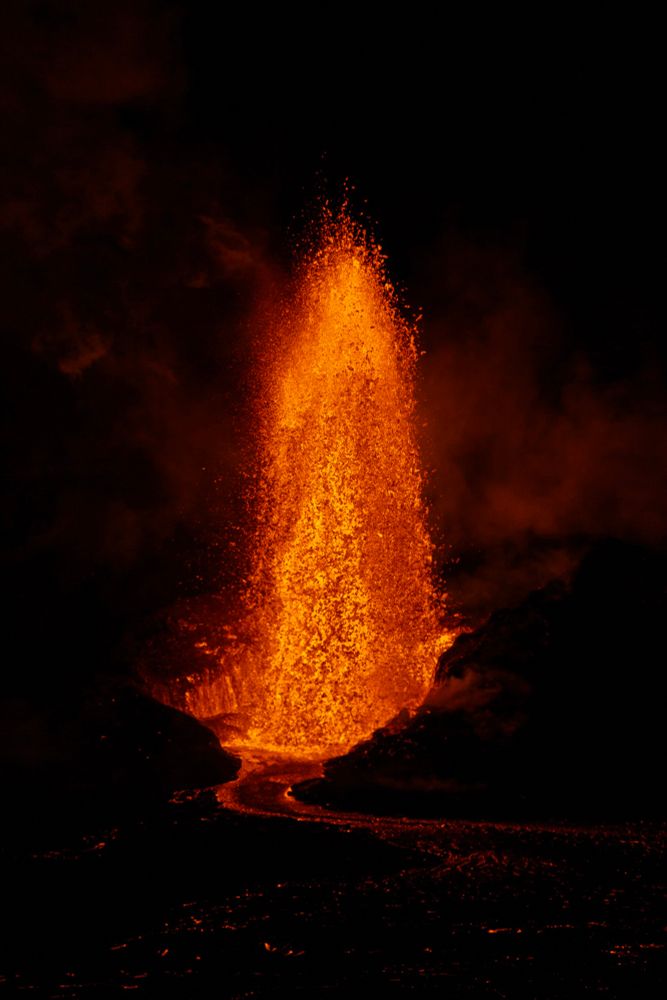 Fountain of lava shooting upwards at night about 400-600 feet. There’s a small river of lava flowing in front of it. 