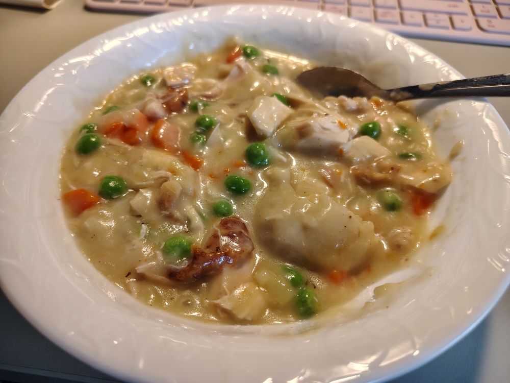 A white bowl filled with creamy chicken and dumplings. You can see bits of chicken, chicken skin, peas, carrots, and dumplings in a thick, creamy sauce