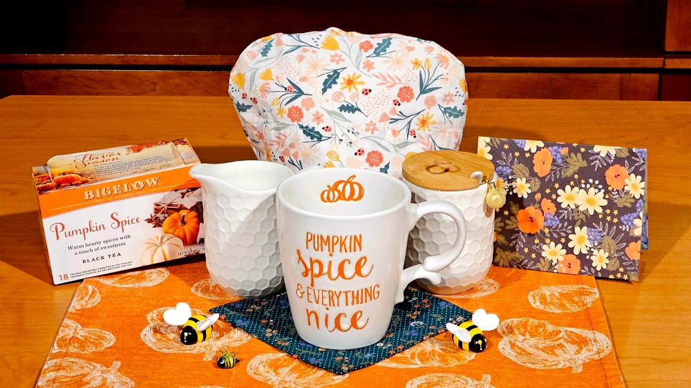 Bright autumn tea tableau with patterns of pumpkins and forest flowers, and bee figurines, and a cream and sugar set that looks like honeycomb, and a mug with the inscription Pumpkin Spice is Everything Nice, and a box of Bigelow Pumpkin Spice black tea. 